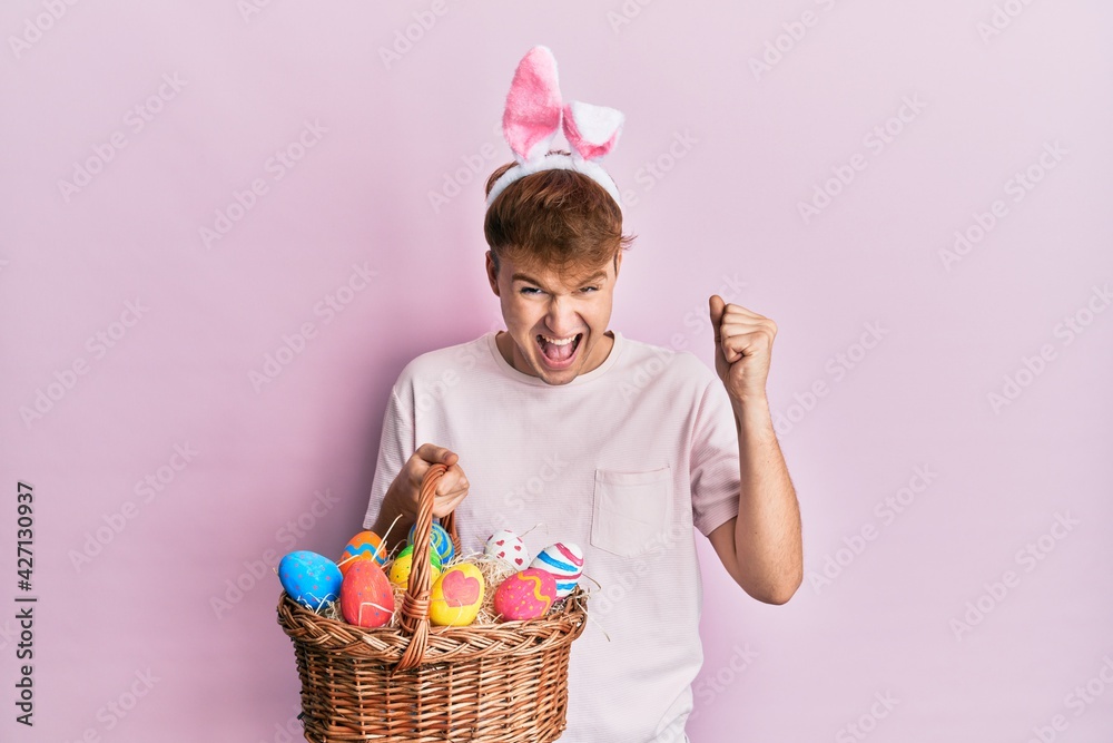 Young caucasian man wearing cute easter bunny ears holding wicker basket with colored eggs screaming proud, celebrating victory and success very excited with raised arms