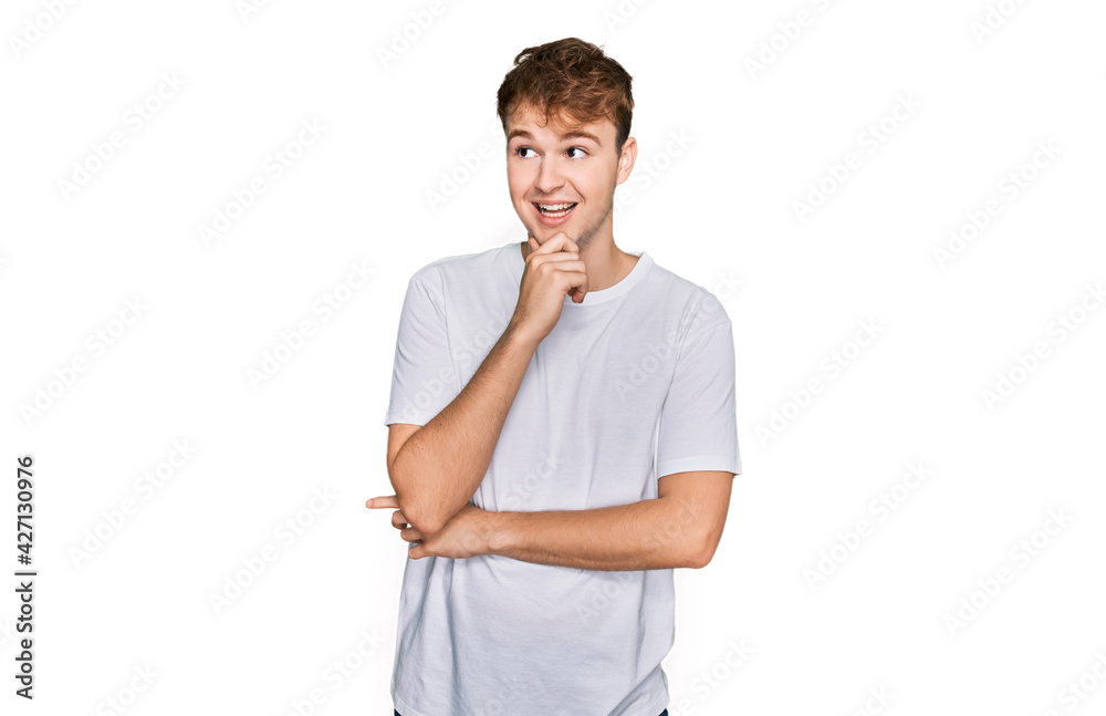 Young caucasian man wearing casual white t shirt with hand on chin thinking about question, pensive expression. smiling with thoughtful face. doubt concept. Young caucasian man wearing casual white t shirt with hand on chin thinking about question, pensive expression. smiling with thoughtful face. doubt concept.