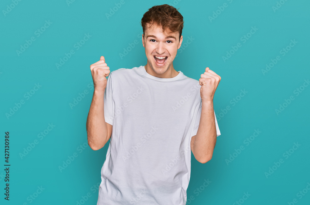 Young caucasian man wearing casual white t shirt celebrating surprised and amazed for success with arms raised and open eyes. winner concept. Young caucasian man wearing casual white t shirt celebrating surprised and amazed for success with arms raised and open eyes. winner concept.
