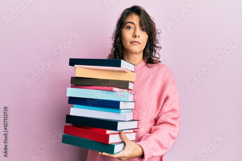 Young hispanic woman holding a pile of books relaxed with serious expression on face. simple and natural looking at the camera.