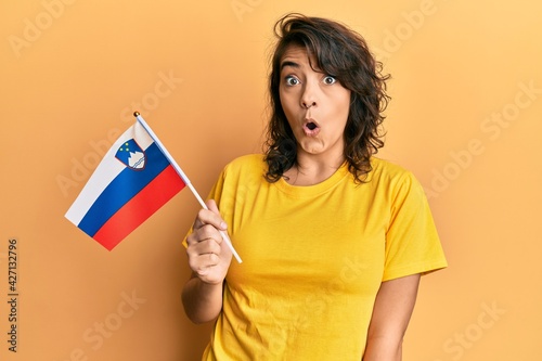 Young hispanic woman holding slovenia flag scared and amazed with open mouth for surprise  disbelief face