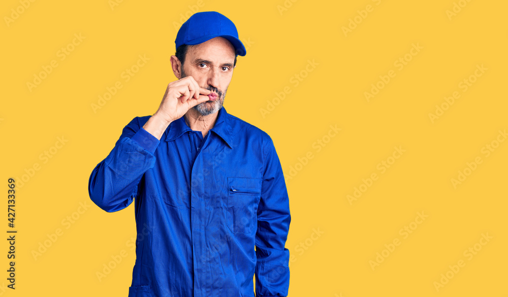 Middle age handsome man wearing mechanic uniform mouth and lips shut as zip with fingers. secret and silent, taboo talking