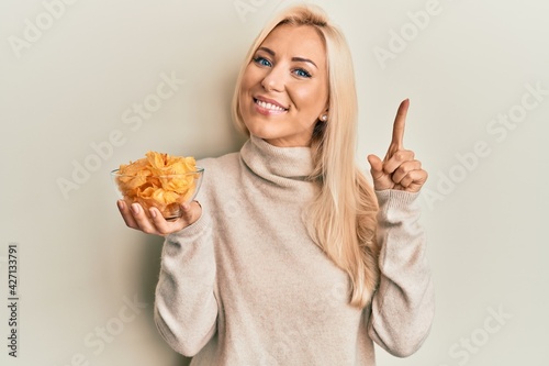 Young blonde woman holding bowl with potato chip smiling with an idea or question pointing finger with happy face  number one