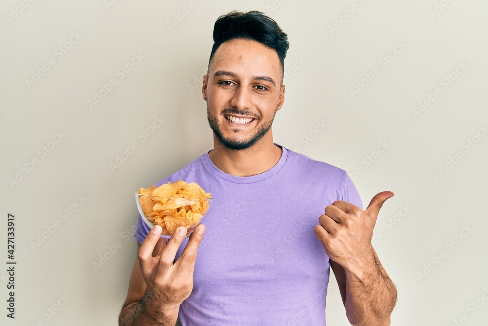 Young arab man holding potato chip pointing thumb up to the side smiling happy with open mouth Young arab man holding potato chip pointing thumb up to the side smiling happy with open mouth