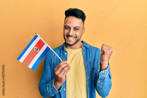 Young arab man holding costa rica flag screaming proud, celebrating victory and success very excited with raised arm