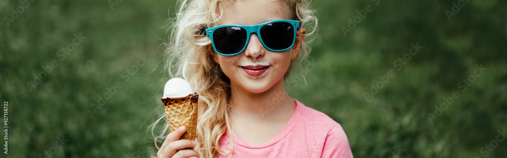 Obraz premium Cute funny adorable girl in sunglasses with dirty nose and moustaches eating ice cream from waffle cone. Happy cool hipster child eating tasty sweet cold summer food outdoor. Web banner header.