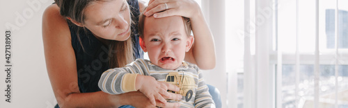  Mother hugging pacifying sad upset crying toddler boy. Family young mom and crying baby sitting on bed at home. Bonding relationship of mom and child baby. Web banner header.