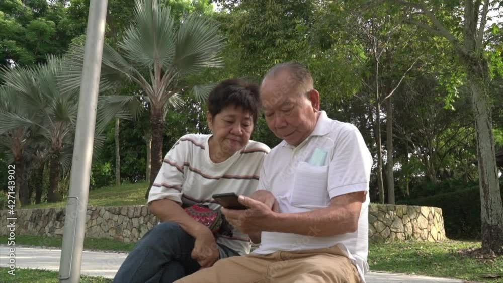 Senior Asian couple sitting on bench in a park, reading and surfing phone together. Senior Asian couple sitting on bench in a park, reading and surfing phone together.