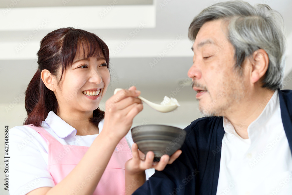 シニア男性に食事の介助をするエプロン姿の若い女性 シニア男性に食事の介助をするエプロン姿の若い女性