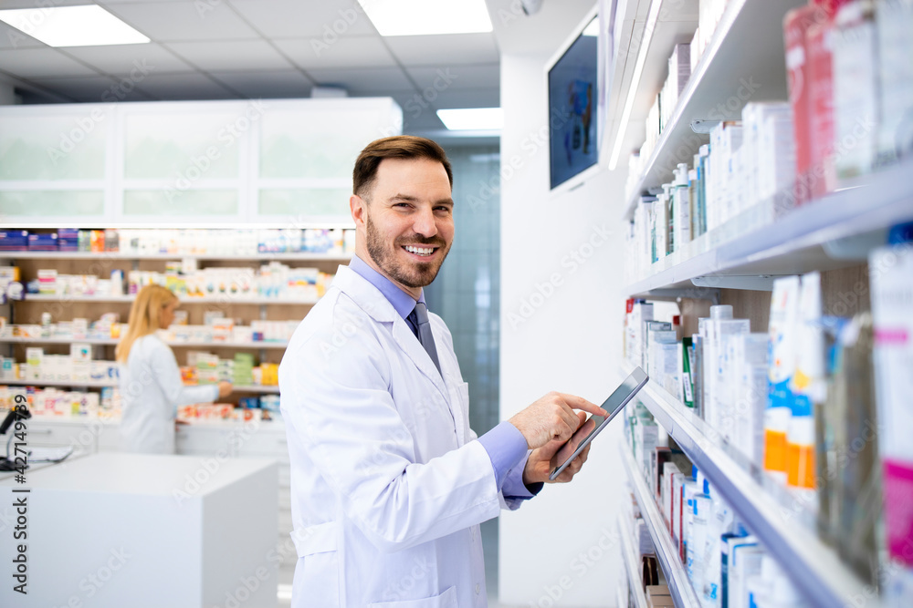 Pharmacist standing by the shelves with medicines and typing on tablet in drug store.