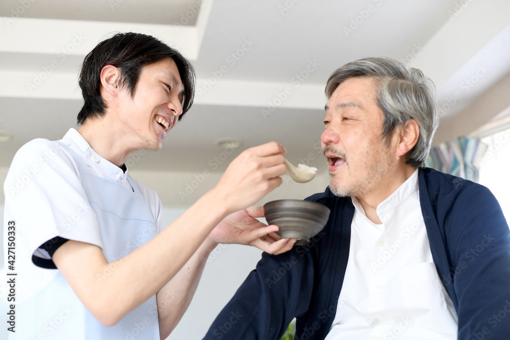 Obraz premium シニア男性に食事の介助をするエプロン姿の若い男性