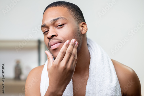 Male grooming. Skin care. Facial treatment. Beard moisturizing. Confident shirtless African man with white towel touching face with hand after cleansing. photo