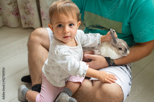 adorable baby sits in dad s arms and strokes a decorative rabbit. domestic animals in a family with children. father shows little child easter bunny