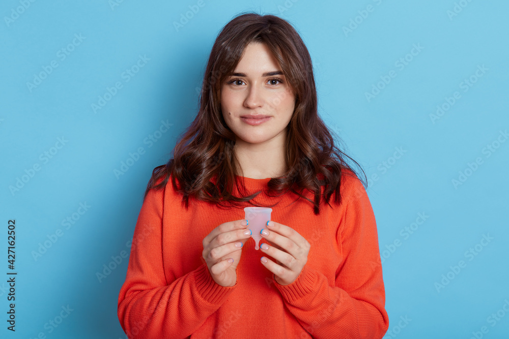 Pleasant looking dark haired girl with calm expression holding menstrual cup in hands, looking directly at camera, choosing alternative variant of hygiene and safety, isolated over blue background. Pleasant looking dark haired girl with calm expression holding menstrual cup in hands, looking directly at camera, choosing alternative variant of hygiene and safety, isolated over blue background.