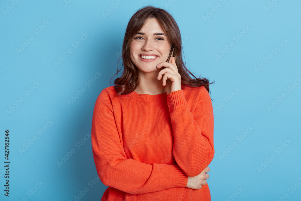 European female with happy facial expression posing with phone near ear, has pleasant conversation, hears excellent news, smiling to camera, dresses in orange jumper, isolated over blue background. European female with happy facial expression posing with phone near ear, has pleasant conversation, hears excellent news, smiling to camera, dresses in orange jumper, isolated over blue background.