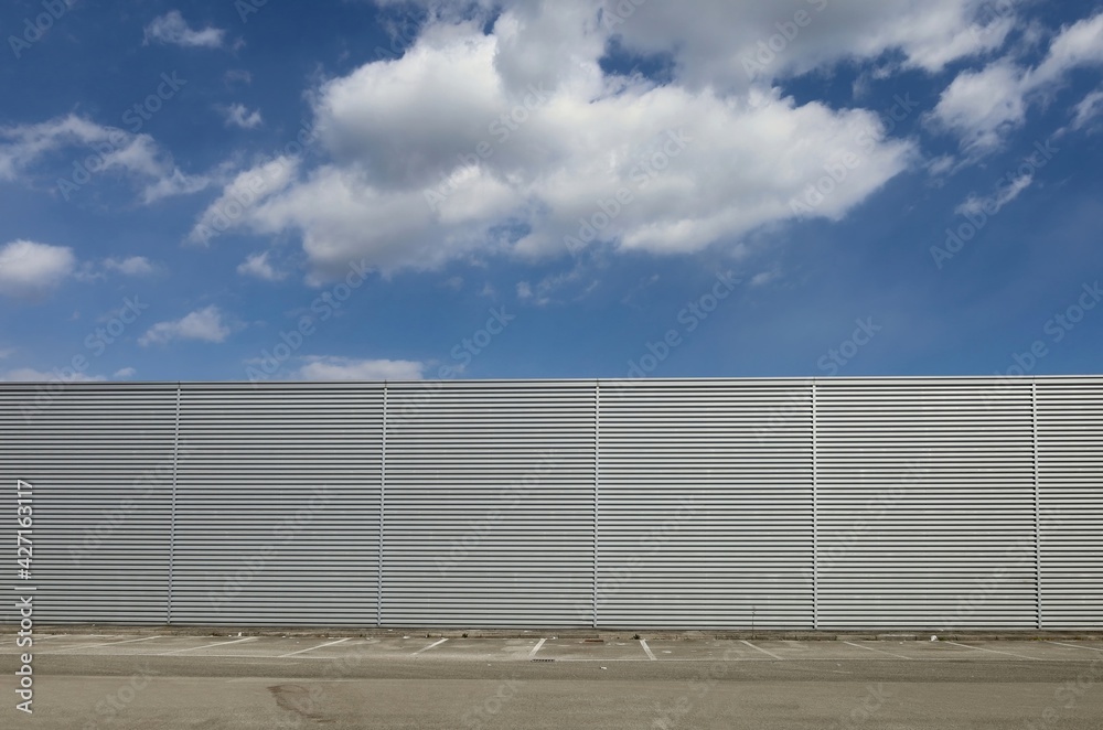 Aluminium cladding wall with horizontal stripes under a blue sky with fluffy clouds. Sidewalk, parking and asphalt road in front. Background for copy space