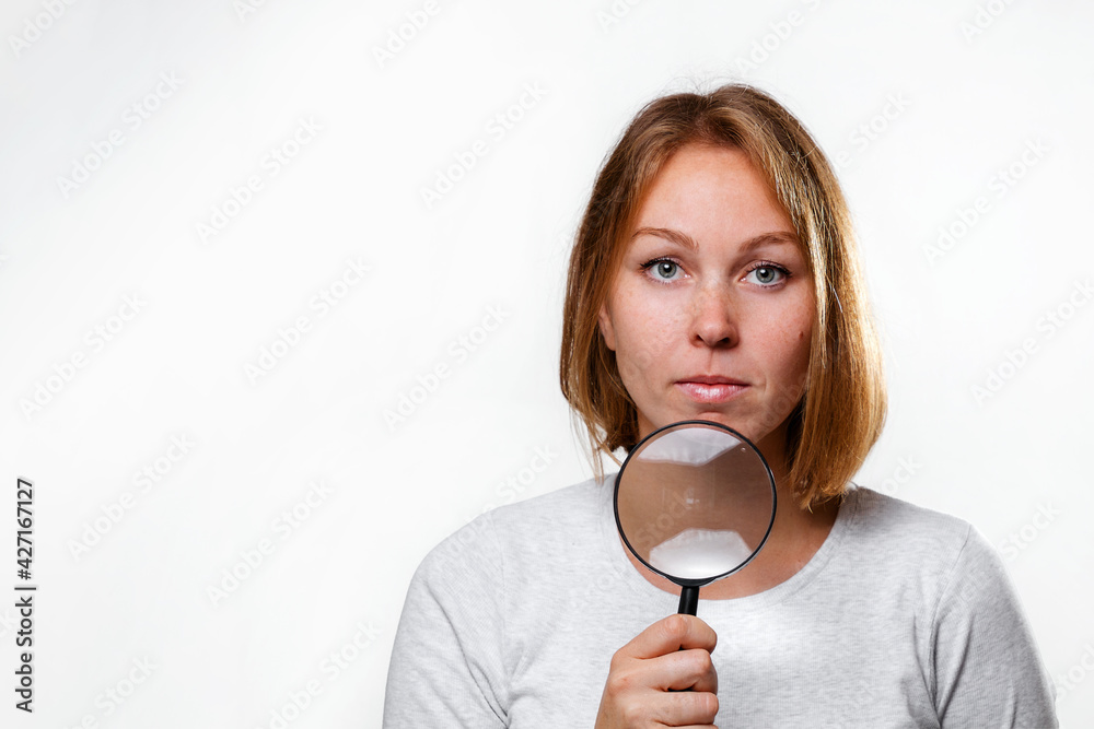 Portrait of a young Caucasian woman holding a magnifying glass to her face. White background. Copy space. The concept of search, research and business Portrait of a young Caucasian woman holding a magnifying glass to her face. White background. Copy space. The concept of search, research and business