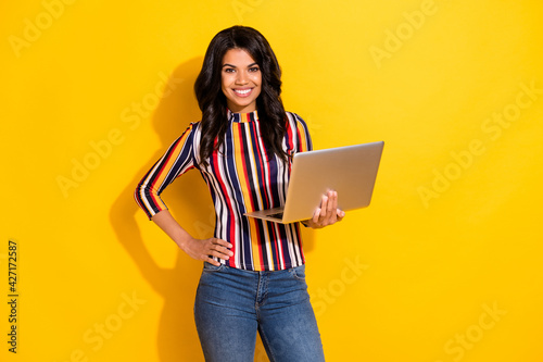 Photo of charming dark skin lady put hand on waist hold laptop toothy smile isolated on yellow color background