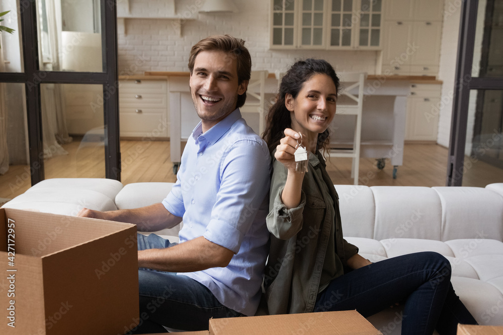 Portrait happy family excited by relocation, purchase new apartment, overjoyed wife showing keys at camera, smiling man and woman sitting on couch, unpacking boxes, mortgage or rent concept Portrait happy family excited by relocation, purchase new apartment, overjoyed wife showing keys at camera, smiling man and woman sitting on couch, unpacking boxes, mortgage or rent concept