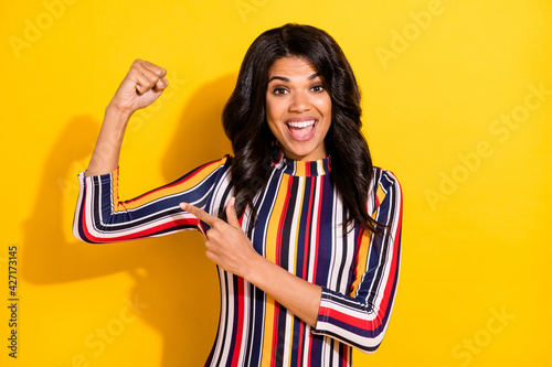 Photo of excited attractive ark skin girl direct fingers hand flexing biceps isolated on yellow color background