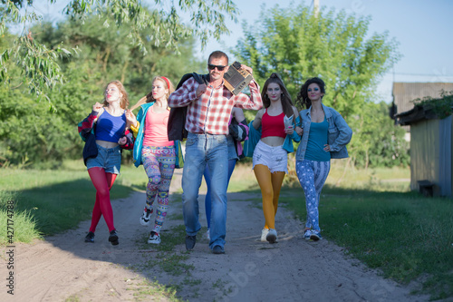 A funny guy with a cassette tape recorder and a group of beautiful girls are walking along the road.