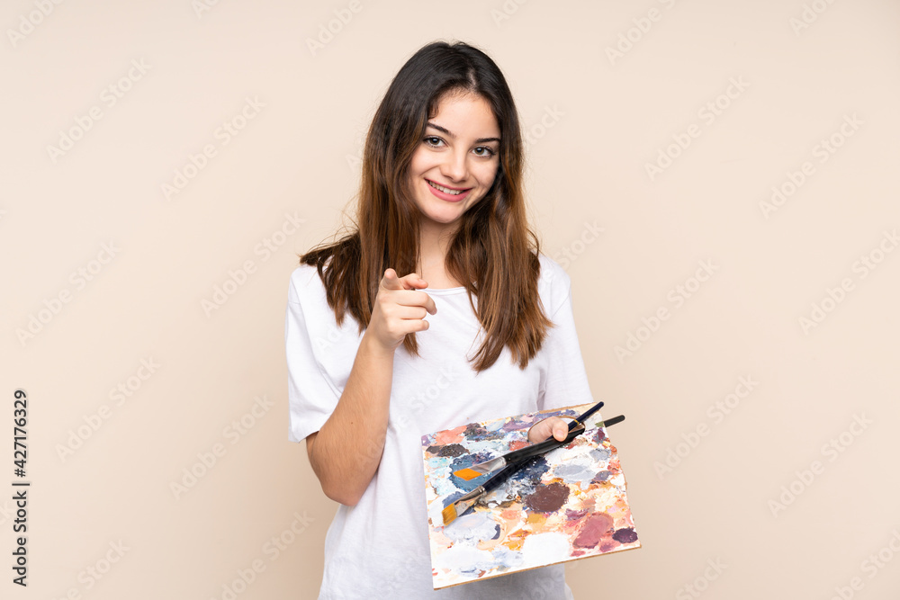 Young artist man holding a palette isolated on beige background points finger at you Young artist man holding a palette isolated on beige background points finger at you