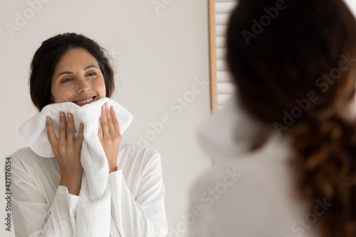 Smiling woman wiping face with white fluffy towel after shower, happy attractive young female wearing bathrobe looking in bathroom mirror, enjoying skincare beauty spa procedure, morning routine