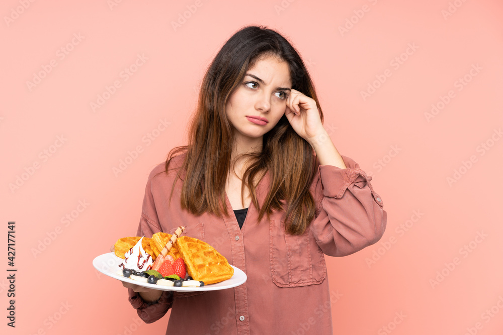 Young caucasian woman holding waffles isolated on pink background having doubts and with confuse face expression