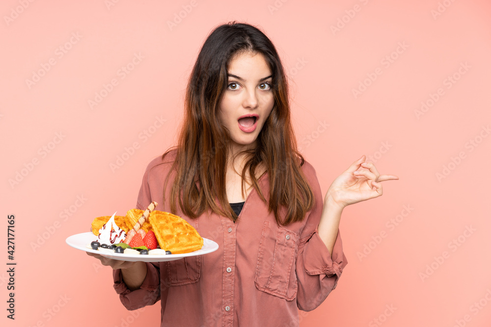 Young caucasian woman holding waffles isolated on pink background surprised and pointing finger to the side