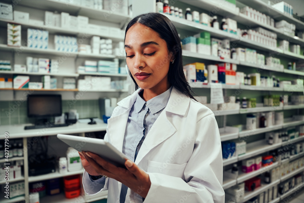 Young female pharmacist working using digital tablet standing in chemist wearing labcoat Young female pharmacist working using digital tablet standing in chemist wearing labcoat