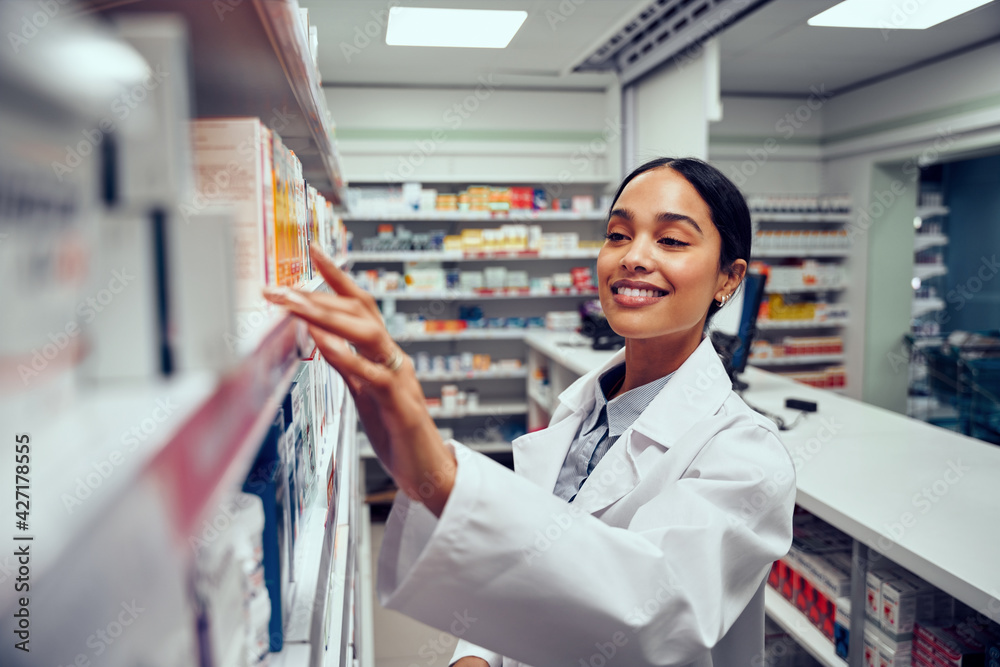 © StratfordProductions - Smiling young female pharmacist wearing labcoat standing behind counter looking for medicine in shelf © StratfordProductions - Smiling young female pharmacist wearing labcoat standing behind counter looking for medicine in shelf