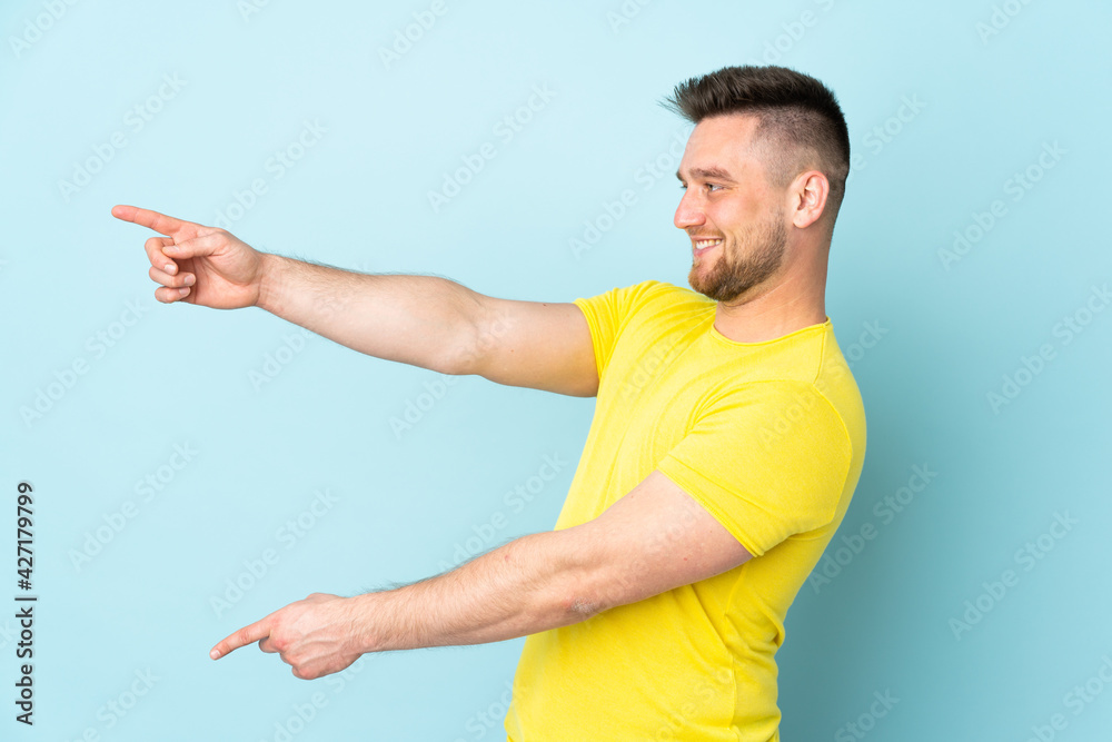 Russian handsome man isolated on blue background pointing finger to the side and presenting a product Russian handsome man isolated on blue background pointing finger to the side and presenting a product