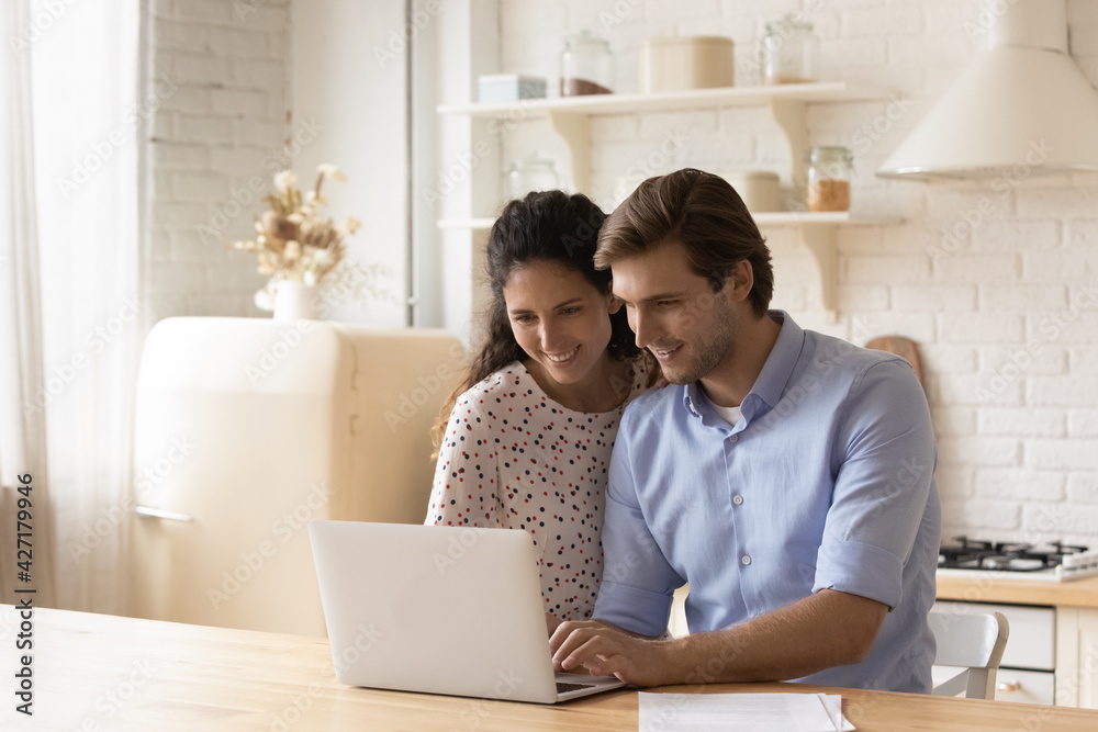 Happy young couple using laptop in modern kitchen together, looking at screen, pleasant family surfing internet, wife and husband shopping or chatting online, browsing internet banking services Happy young couple using laptop in modern kitchen together, looking at screen, pleasant family surfing internet, wife and husband shopping or chatting online, browsing internet banking services