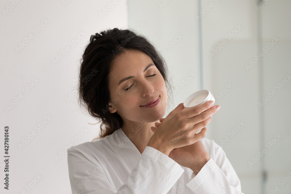 Close up happy beautiful woman applying moisturizing hand cream, smiling attractive young female in bathrobe holding plastic jar with balm or lotion, enjoying skincare spa procedure, beauty treatment Close up happy beautiful woman applying moisturizing hand cream, smiling attractive young female in bathrobe holding plastic jar with balm or lotion, enjoying skincare spa procedure, beauty treatment