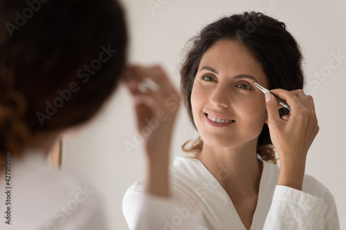 Close up smiling woman in white bathrobe plucking thick eyebrows with tweezers, looking in mirror, attractive young female correcting shape, beauty procedure and make up, standing in bathroom