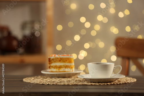Hot drink in cup and piece of cake on wooden table against blurred lights indoors