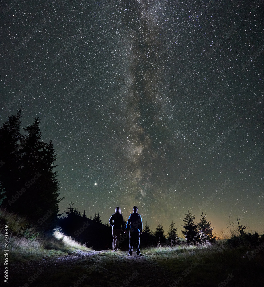 Fototapeta premium Picturesque view of night starry sky over mountain valley with couple travelers. Young woman and man holding hands while standing on mountain road under beautiful sky with stars and Milky Way.