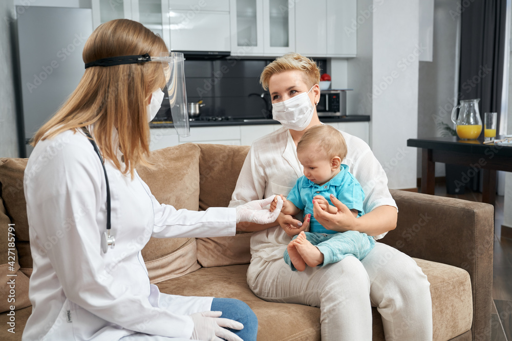 Obraz premium Competent doctor in protective mask and gloves visiting her little patient at home. Young mother sitting on couch with cute toddler i on hands. Medical treatment.