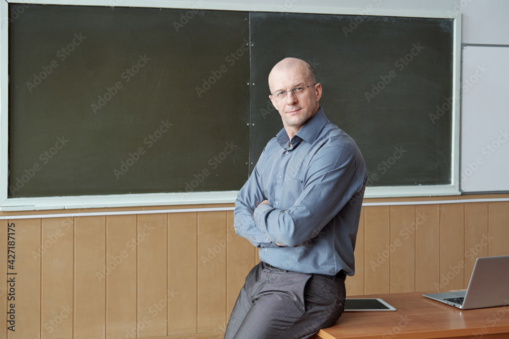 Bald professor of university in smart casualwear sitting on desk against blackboard while crossing arms by chest and looking at you Bald professor of university in smart casualwear sitting on desk against blackboard while crossing arms by chest and looking at you