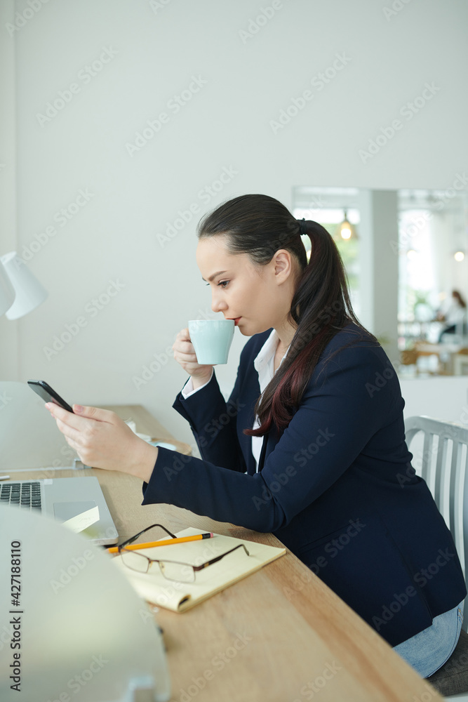 Beautiful young businesswoman enjoying cup of coffee in cafe and reading news or text messages on smartphone Beautiful young businesswoman enjoying cup of coffee in cafe and reading news or text messages on smartphone