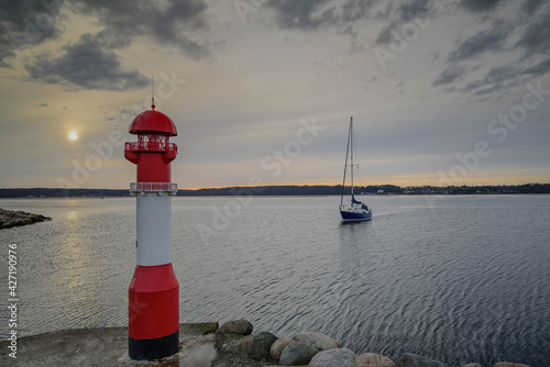 The red pier light with sailing boot enter in marina (dock, basin with moorings and supplies for yachts ans small boots). Red pier light at the entrance to the marina Fahrensodde by sunset, Flensburg. photo