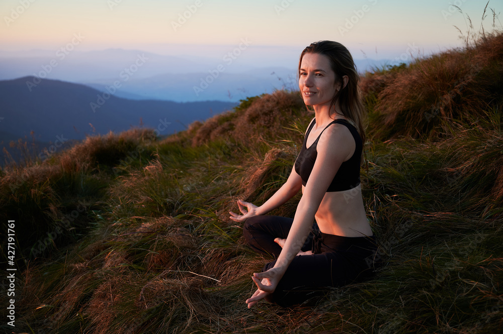 Side view of brunette woman practicing yoga on nature with beautiful landscape on background. Concept of meditation.