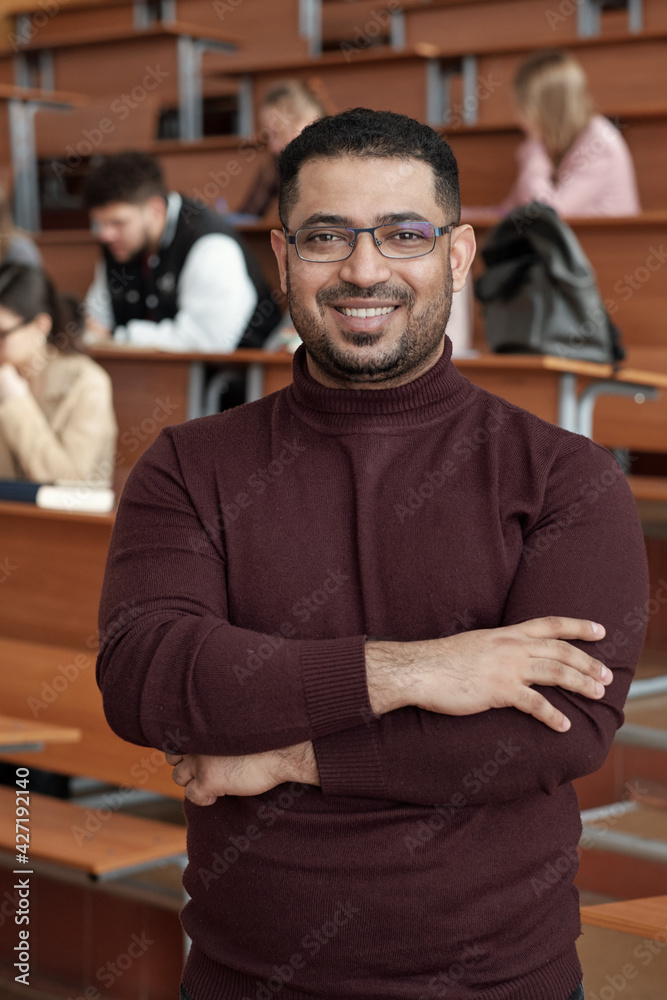 Young successful male teacher crossing his arms by chest while standing in front of camera against students sitting by desks in lecture hall Young successful male teacher crossing his arms by chest while standing in front of camera against students sitting by desks in lecture hall