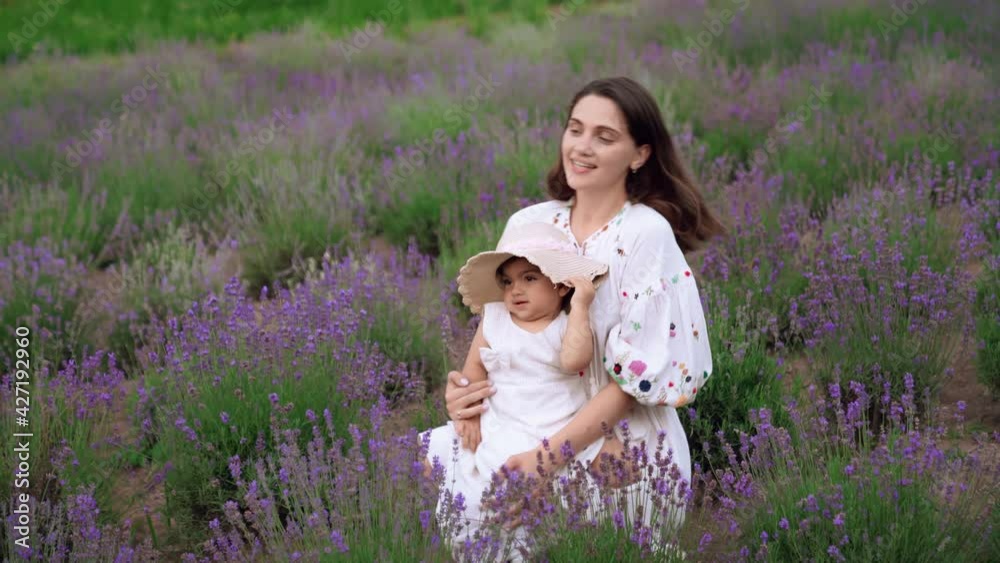 Motion of caucasian mother enjoying time with little cute baby daughter wearing straw hat and dress, kissing and hugging daughter. Young woman showing love, sitting in blooming lavender field. Motion of caucasian mother enjoying time with little cute baby daughter wearing straw hat and dress, kissing and hugging daughter. Young woman showing love, sitting in blooming lavender field.