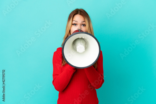 Teenager blonde girl over isolated blue background shouting through a megaphone to announce something