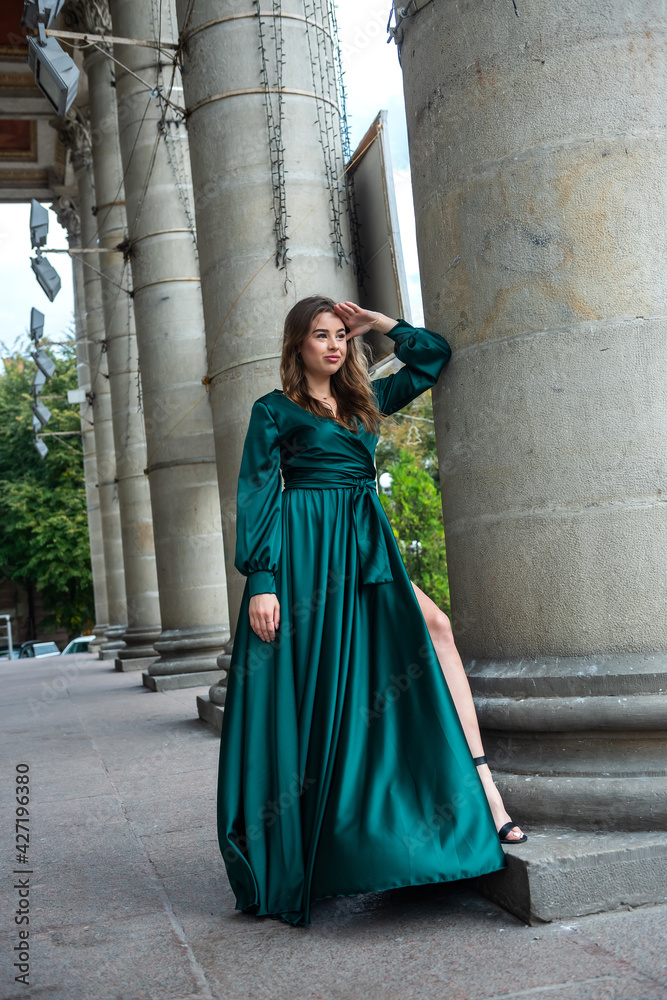 Naklejka premium young elegant woman relaxing and posing for camera near columns in green dress