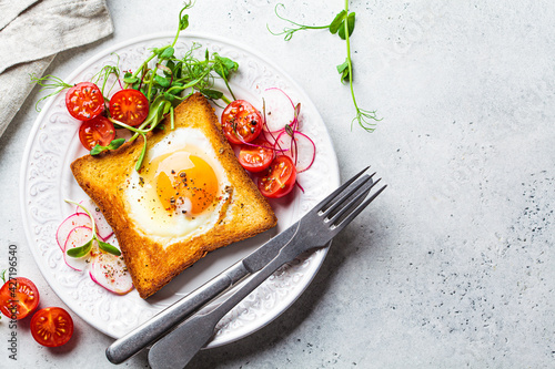 Fried egg in bread toast with tomatoes, radishes and sprouts on white plate, gray background, top view. Healthy breakfast concept.