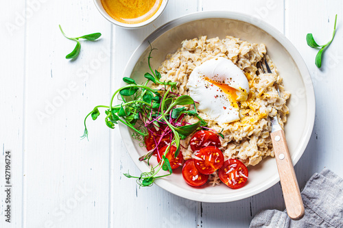 Savory oatmeal with poached egg, tomatoes, cheese and sprouts in white bowl, top view. Healthy breakfast concept.