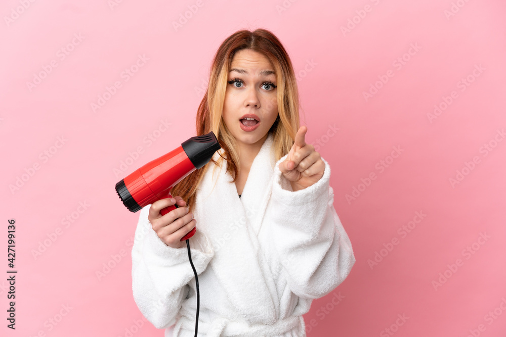 Teenager blonde girl holding a hairdryer over isolated pink background surprised and pointing front Teenager blonde girl holding a hairdryer over isolated pink background surprised and pointing front