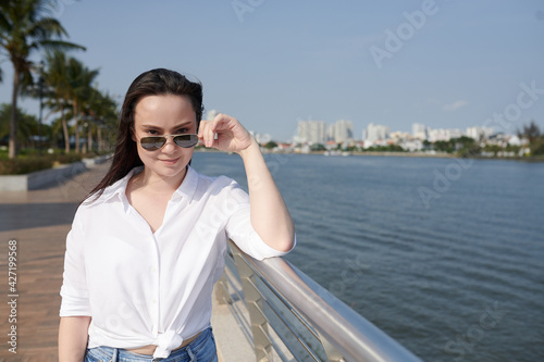 Portrait of young beautiful woman taking off sunglasses and smiling at camera when standing on river bank © DragonImages